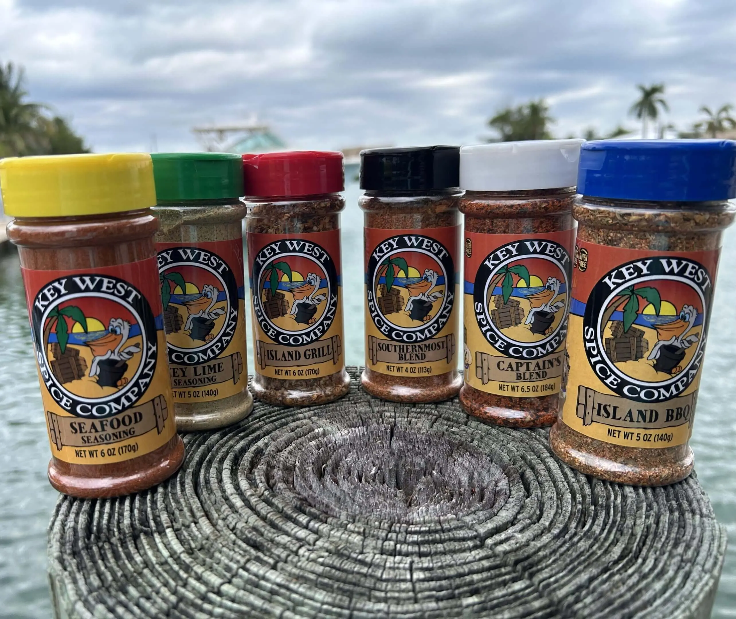 A lineup of seven colorful spice bottles on a woven surface under a cloudy sky.